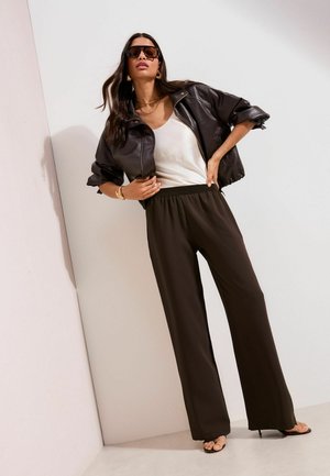 Nő sötétbarna bőrkabátban, fehér felsőben, fekete bőszárú nadrágban, napszemüvegben, arany ékszerekkel, fehér fal előtt állva.