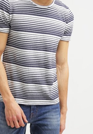 Hombre con camiseta de manga corta con rayas horizontales azul marino y blancas y vaqueros azules, de pie frente a un fondo blanco liso.