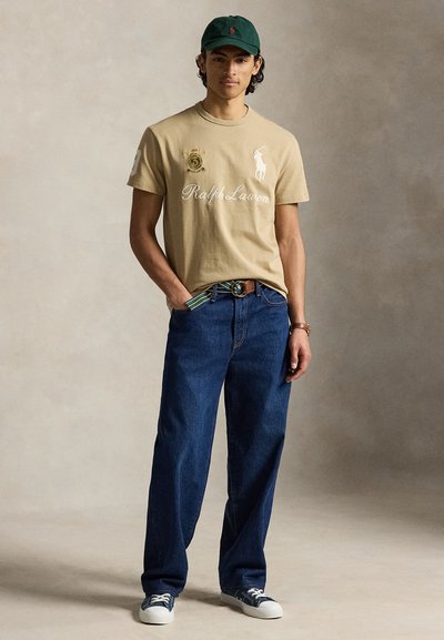 Νέος άνδρας που φοράει μπεζ μπλούζα Ralph Lauren, μπλε τζιν, ναυτικές αθλητικές παπούτσια, σκούρο πράσινο καπέλο και πολύχρωμη ζώνη, στέκεται με το ένα χέρι στην τσέπη.