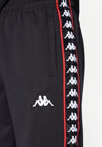 Zwarte joggerbroek met een elastische tailleband, voorzien van rode strepen aan de zijkant en een herhalend wit Kappa-logo patroon. Zachte stoftextuur.