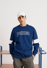 Navy sweatshirt with white accents and "puma" logo on the front, paired with a white cap. Model stands against a minimalist background.