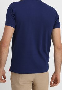 Marinblå pikétröja i bomull. Kort ärm, rund halsringning, sidoveck med röda accenter i fållen. Slät yta.