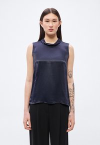 Tmavomodrý saténový top bez rukávov s cowl výstrihom a hladkou, lesklou textúrou. Lem je jemne zakrivený, čo dopĺňa široké čierne nohavice s rovnými nohavicami.