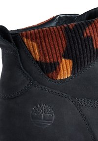 Černá kožená bota s oranžovým, hnědým a černým vzorovaným kordovým vrškem a vyraženým logem stromu poblíž paty.
