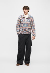 Νέος άνδρας που φοράει πολύχρωμο πουλόβερ, λευκό πουκάμισο με γιακά, φαρδιά μαύρη παντελόνα cargo και καφέ παπούτσια, στέκεται μπροστά σε απλό φόντο.