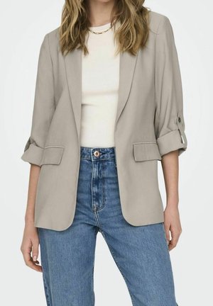 Femme portant un blazer beige retroussé, une chemise blanche et un jean bleu taille haute, montrée des épaules aux hanches.