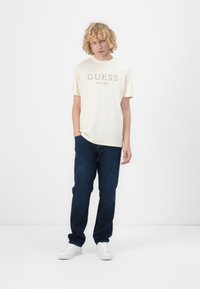 Majica od pamučnog materijala boje kreme s grafikom "GUESS EST. 1981", uparena s tamnoplavim trapericama i bijelim tenisicama.
