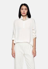 Weiße Bluse mit langen Ärmeln, hohem Kragen, Knopfleiste und lockerer Passform. Weicher Stoff mit leichter Textur. Kombiniert mit passenden Hosen.