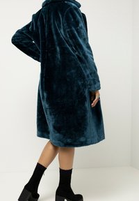 Dlouhý tyrkysový kabát z umělé kožešiny s límcem, s jemnou texturou a bočními kapsami, nošený přes černé kotníkové boty.