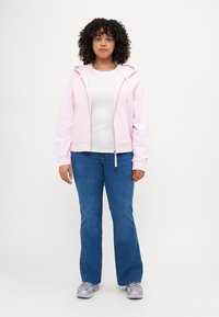 Lichtroze zip-up hoodie met een ruime capuchon, die over een wit shirt is gedragen. Gecombineerd met blauwe flared jeans en grijze sportschoenen.