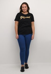 Svart kortärmad T-shirt med gyllene kursivtext som säger "Different", i kombination med blå tajta jeans och svarta loafers, stående mot en neutral bakgrund.