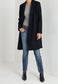 Person klädd i en svart knälång kappa över en grå skjorta, blå jeans och svarta ankelstövlar, stående med en hand i fickan.