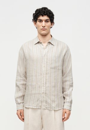 Jeune homme aux cheveux bouclés et foncés, portant une chemise beige rayée à manches longues boutonnée et un pantalon clair, debout devant un fond uni.