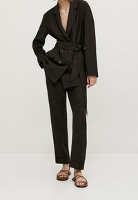 Blazer marron foncé ajusté avec un design cache-cœur, boutons croisés et pantalon assorti. Porté avec des sandales marron à brides.