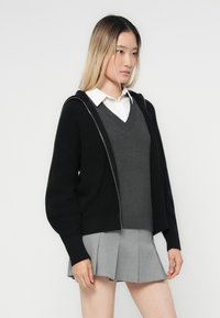 Zwarte gebreide cardigan met een rits aan de voorkant, gedragen over een grijze v-hals trui en een witte blouse. Vergezeld door een grijze geplisseerde rok.