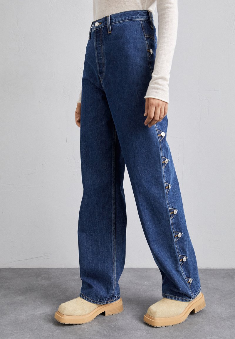 Jeans iz jeansa v temno modri barvi z visokim pasom, ravnimi hlačnicami in gumbnimi detajli ob strani. V kombinaciji z bež platformasto obutev.