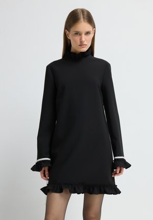 Zwarte jurk met lange mouwen, gerimpelde manchetten en een gerimpelde zoom. Heeft een hoge halslijn en een gladde textuur. Accenten in het wit.