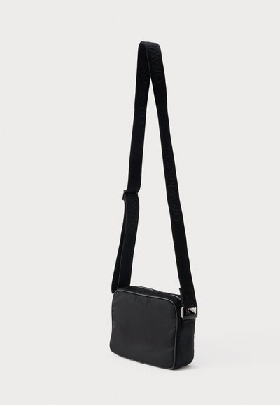 Μαύρη ορθογώνια τσάντα χιαστί με ρυθμιζόμενο λουρί και διακριτικό ανάγλυφο λογότυπο στο λουρί, μονόχρωμο φόντο.