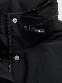 Sort jakke med høj krave, tekstureret stof, sølvtrykknap og sort lynlås med en mærke-emballeret trækstrop.