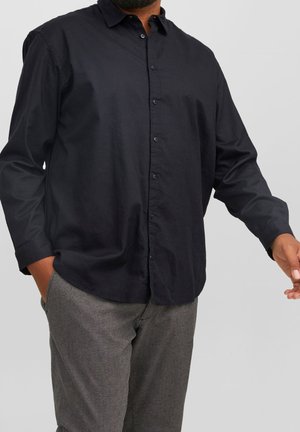 Mann trägt ein schwarzes Hemd mit Knöpfen und graue strukturierte Hose, mit einer Hand in der Tasche und der anderen leicht nach vorne ausgestreckt.