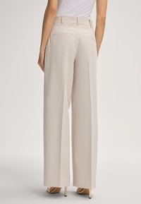 Donna che indossa pantaloni beige a vita alta e gamba larga con tasche a filetto posteriori e sandali beige con tacco alto, in piedi contro uno sfondo semplice.