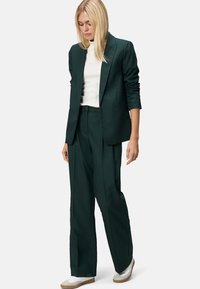 Blazer ajusté vert foncé et pantalon large, associés à un col roulé blanc et des baskets blanches, mettant en avant un look professionnel élégant.