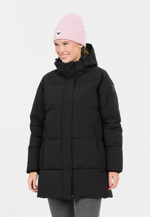 Zwarte pufferjack met een hoge kraag, ritszakken en een matte afwerking. Een roze gebreide beanie met een logo wordt op het hoofd gedragen.