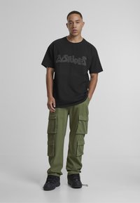 Must musta suur hall graafikuga, mida on kombineeritud oliivroheliste cargo-pükste ja mustade spordijalanõudega. Pükstel on mitmeid taskuid.