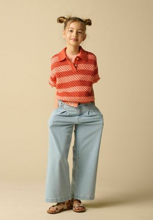 Jong meisje met dubbele knotkapsel draagt een rood gestreept gebreid topje, wijde lichtblauwe jeans en sandalen, staand tegen een beige achtergrond.