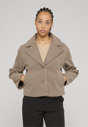 Kvinne med fletter i håret har på seg en beige og brun jakke med fiskebensmønster over en beige topp og svarte bukser, står med hendene i jakkelommene.