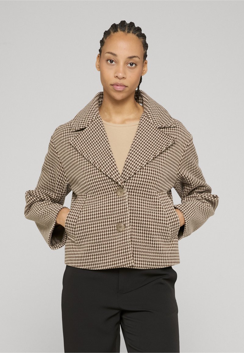 Femme aux cheveux tressés porte une veste beige et marron à motif pied-de-poule sur un haut beige et un pantalon noir, debout les mains dans les poches de la veste.