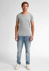 Tricou gri cu gât în V, realizat din material moale, asortat cu blugi din denim deschis la culoare și pantofi sport albi. Modelul stă drept pe un fundal neutru.