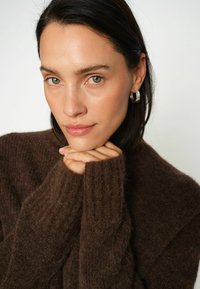 Žena s tmavými vlasy a zelenýma očima, opírající bradu o ruce, nosící stříbrné kruhové náušnice a hnědý chlupatý svetr.