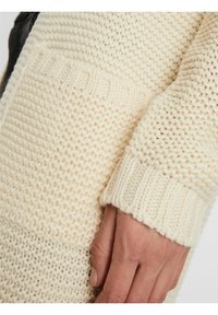 Gruby kardigan dzianinowy w kremowym kolorze z prążkowanymi mankietami i teksturowanym wzorem; widoczna ręka trzymająca tkaninę, ukazująca szczegóły i grubość.
