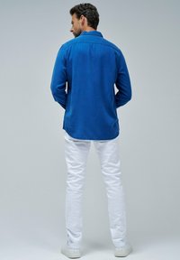 Blaues Langarmhemd aus weichem Stoff, mit einem Button-Down-Kragen und abgerundetem Saum, kombiniert mit weißen Slim-Fit-Hosen und weißen Sneakers.