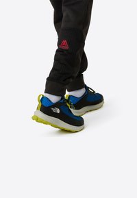 Blå og svarte trening sko med strukturert overdel, lyse gule detaljer og robust yttersåle, kombinert med svarte joggebukser og hvite sokker.