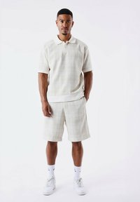 Lichtbeige polo shirt met korte mouwen en witte accenten, gecombineerd met bijpassende shorts. Gelaagd stof met een subtiel geruit patroon. Witte sneakers.