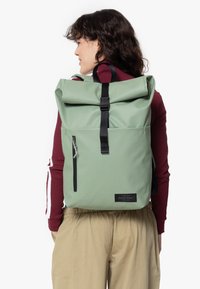 Zaino verde a roll top realizzato in tessuto resistente, dotato di una tasca frontale con zip, spallacci neri e una patch con logo sulla parte inferiore.