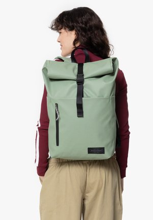 Mochila verde tipo roll-top hecha de tela duradera, con un bolsillo frontal con cremallera, correas negras y un parche con el logo en la base.