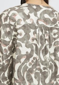 Vue arrière d'une personne portant une chemise ample à manches longues avec des motifs abstraits en spirale dans des tons de gris, blanc et rose clair.