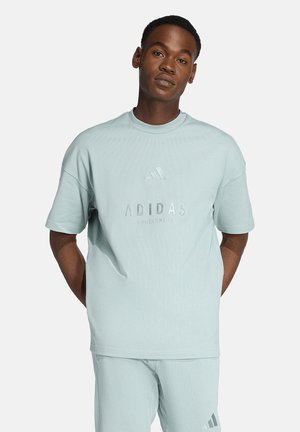 Mann steht mit den Händen hinter dem Rücken und trägt ein hellblaues Adidas-Sport-T-Shirt sowie passende Shorts vor einfarbigem Hintergrund.