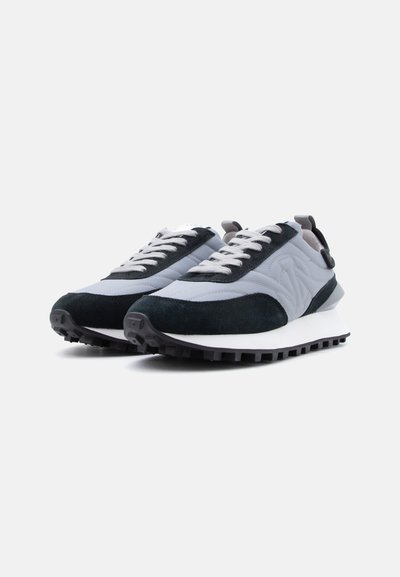 Χαμηλού προφίλ γκρι και μαύρα αθλητικά παπούτσια με λευκά κορδόνια και χοντρή λευκή σόλα με βαθιά πέλματα, με λεπτομέρεια λογοτύπου στο πλάι, εμφανιζόμενα σε τριών τετάρτων όψη.