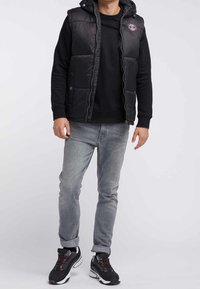 Chaleco negro acolchado con capucha y bolsillos frontales, usado sobre una sudadera negra, combinado con jeans gris claro y zapatillas negras.