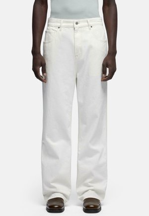 Mand iført løse hvide jeans og mørkebrune sko, stående med armene afslappet ned langs siderne mod en ensfarvet baggrund.