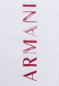Camiseta de algodón blanca con un logo vertical "ARMANI" en letras rojo profundo con brillo, acompañado de formas y líneas geométricas.