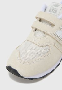 Bézs sportcipő hálós és velúr anyagokból, tépőzáras pántokkal, texturált felsőrésszel és fekete gumitalppal. Klasszikus New Balance dizájn.