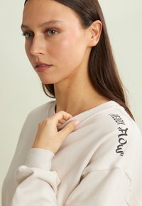 Felpa color beige chiaro con scollo rotondo, polsini a coste e la parola "flow" in nero sulla spalla sinistra. Texture del tessuto morbida e evidente.