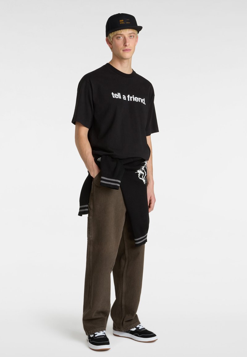 T-shirt en coton noir avec un texte blanc indiquant "dis à un ami", associé à un pantalon ample marron et un pull noir noué autour de la taille. Casquette noire et baskets.