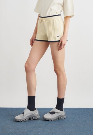 Modelo femenina vistiendo pantalones cortos de punto Adidas beige con ribete negro, calcetas negras y zapatos deportivos grises tipo slip-on, de pie sobre una alfombra azul.