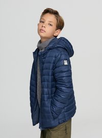 Giacca imbottita blu navy con cuciture orizzontali, cappuccio e logo sulla manica sinistra. Indossata sopra un dolcevita grigio.